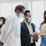 Wearing masks because of the coronavirus outbreak, three doctors confer together. In the background, a female patient and her doctor also wear protective masks.